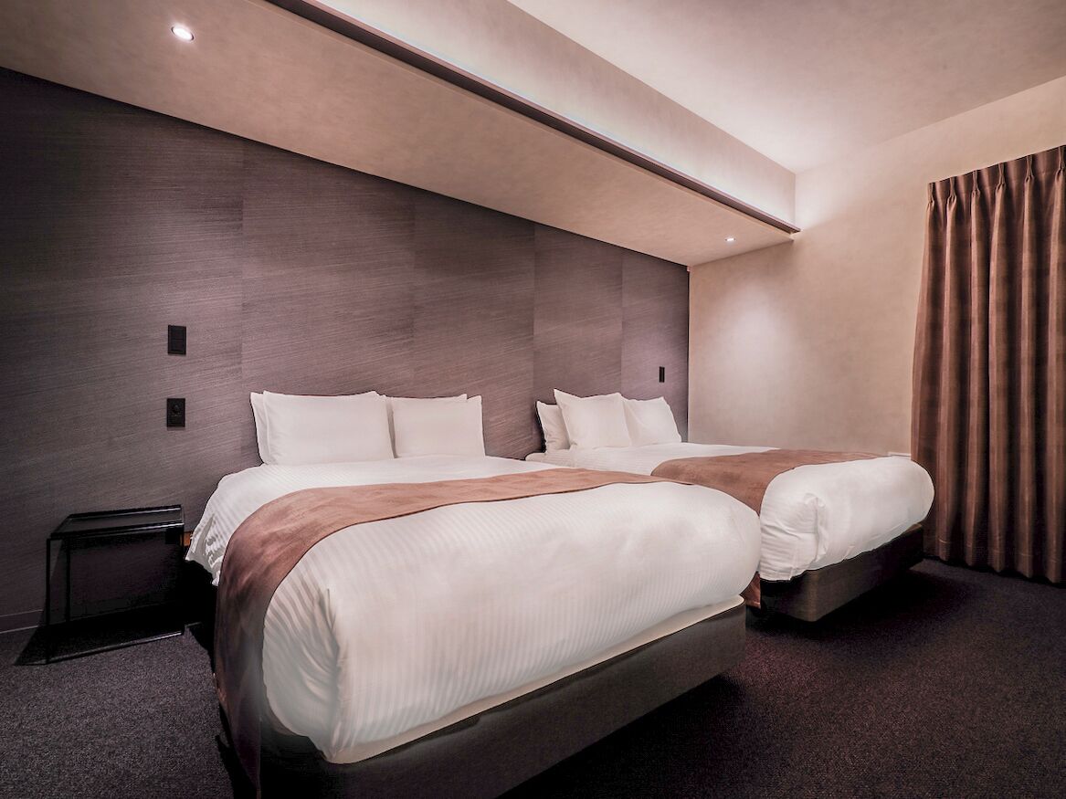 Ein modernes Hotelzimmer mit zwei Betten, einem dunklen Holz-Kopfteil und einem großen Fenster mit Vorhängen.