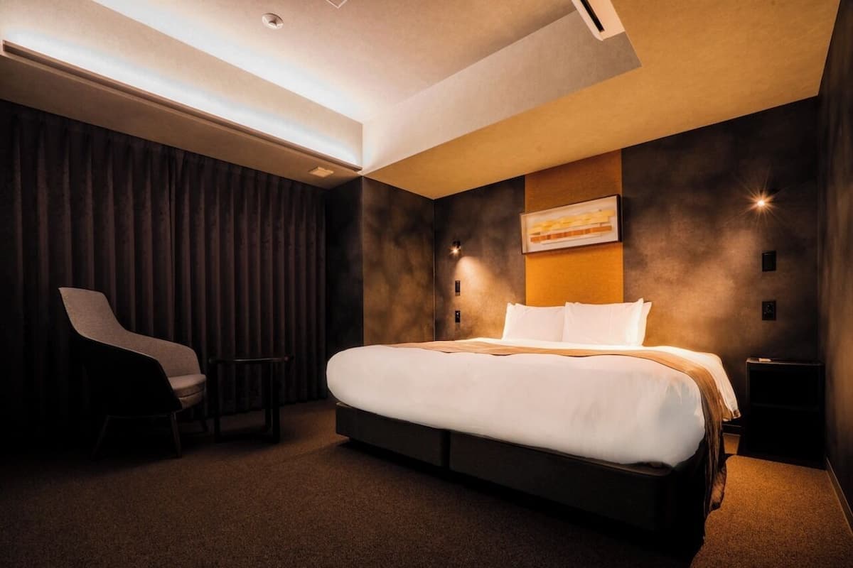 Una habitación de hotel moderna con una cama grande, una silla, una mesita pequeña y una lámpara fijada a la pared.