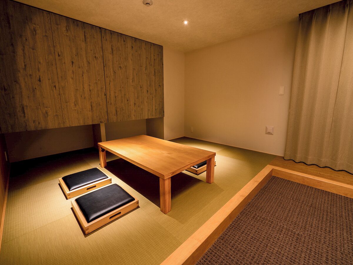 A room with tatami flooring, a low wooden table, two tatami mats with cushions, and a wooden partition wall.
