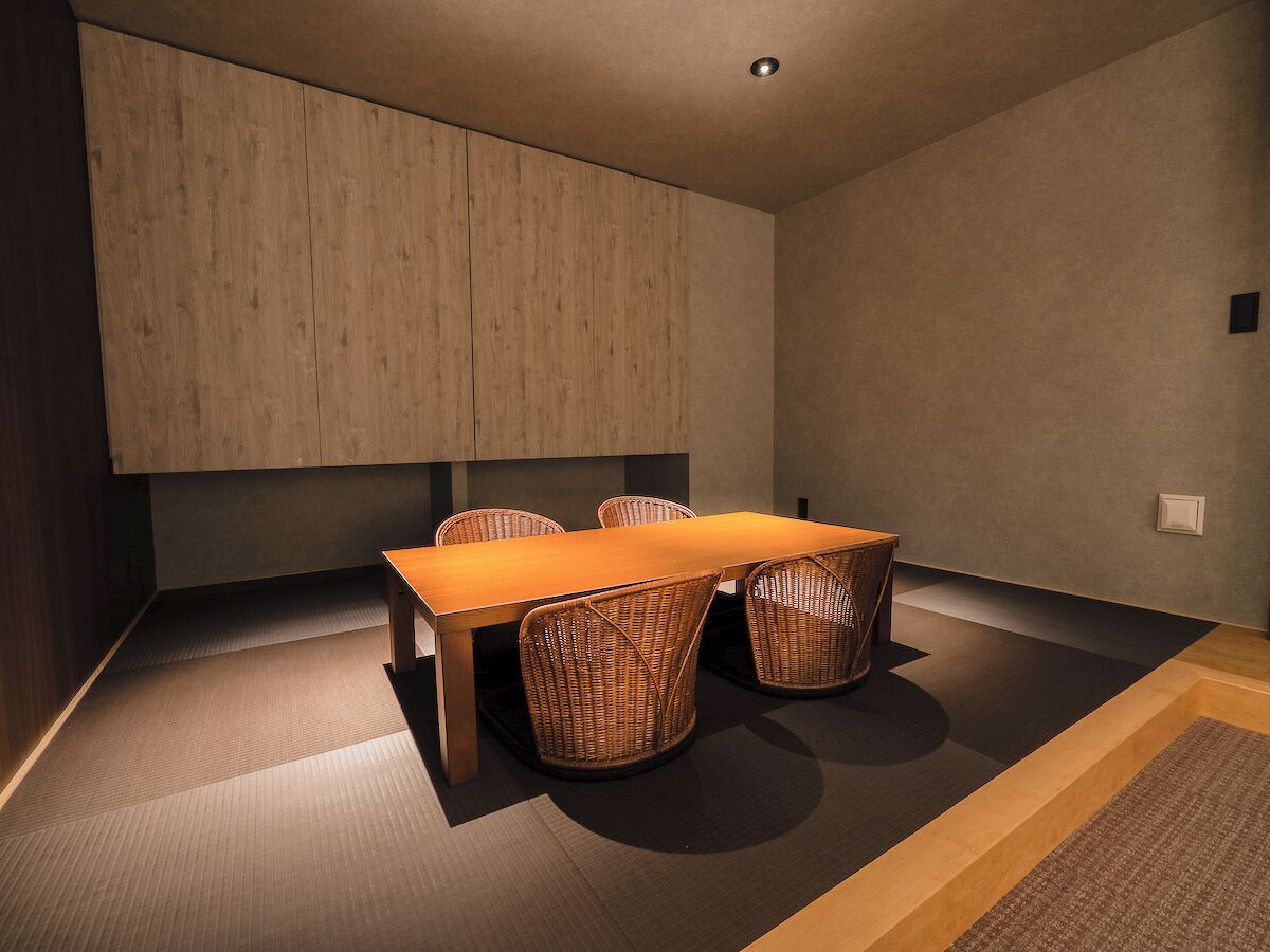 A modern room with a wooden table and wicker chairs, a neutral wall, and a carpeted floor.
