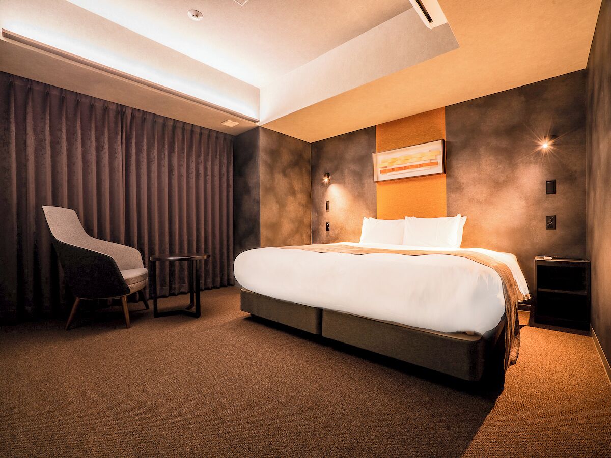 A modern hotel room with a large bed, a chair, a small table, and a wall-mounted light.