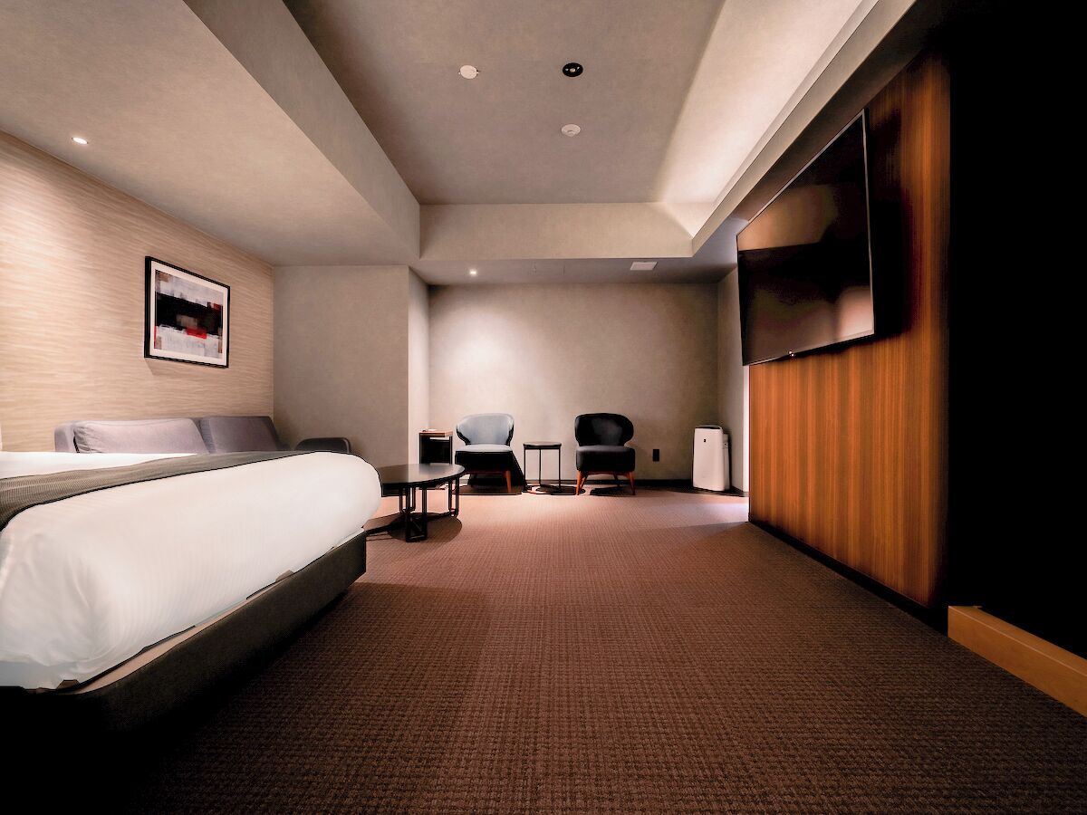 Ein Hotelzimmer mit Bett, zwei Sesseln, einem kleinen Tisch, einem Fernseher und einer Wand aus Holzpaneelen.