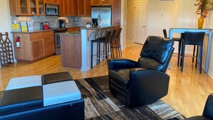 A modern kitchen with wooden cabinets, a central island, and bar stools. There is a black leather chair and a small table with chairs in the living area. - Ultra Modern Condo located in the Heart of Yachats! Walk to everything! (Yachats)