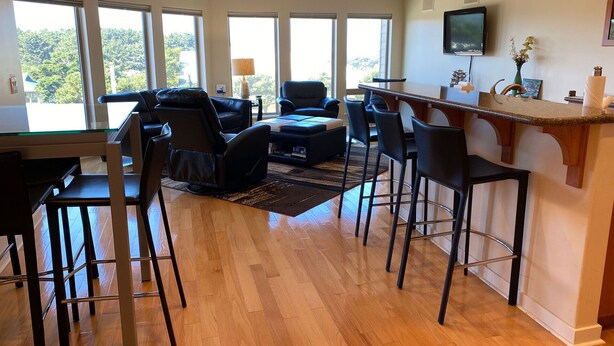 A modern living room with a dining area, large windows, a flat-screen TV, and a kitchen counter. - Ultra Modern Condo located in the Heart of Yachats! Walk to everything! (Yachats)