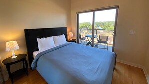 A bedroom with a large bed, two bedside tables with lamps, a balcony with a table and chairs, and a view of greenery outside. - Ultra Modern Condo located in the Heart of Yachats! Walk to everything! (Yachats)