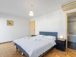 A neatly made bed with white linens, a blue headboard, and bedside lamps. There is a wooden floor and a framed picture on the wall.