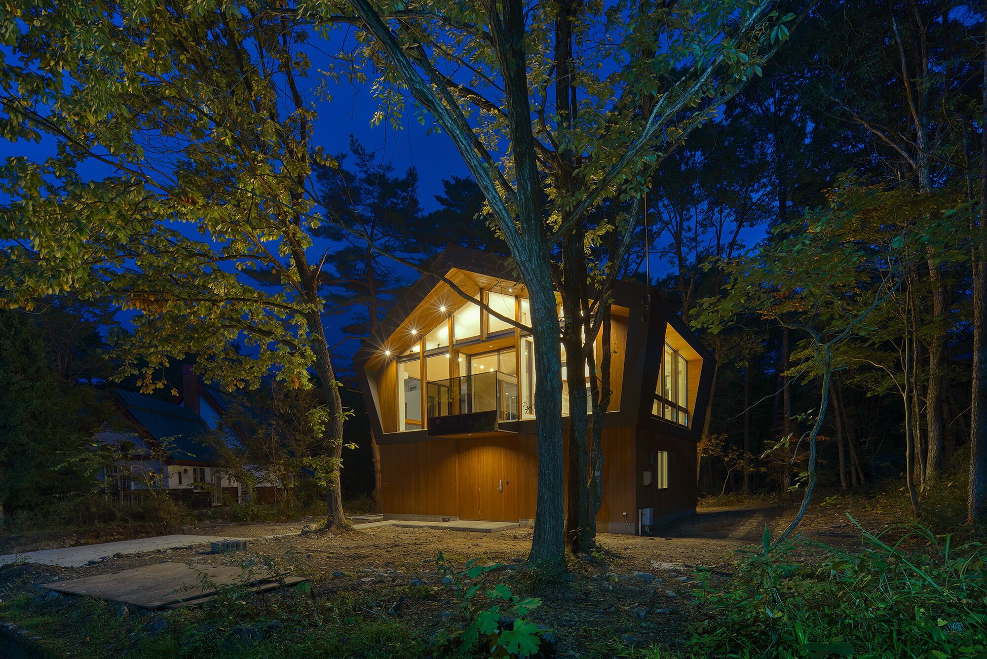 A modern, wooden cabin with large windows, surrounded by trees and a dirt path.