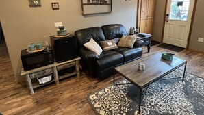 A living room with a black leather sofa, a coffee table, a side table, a microwave, and a refrigerator. There is a door with a wooden frame and a window with a view of the outside. - Wounded Knee Ranch at the Red Hills. Full separate and private suite with deck (Newberg)