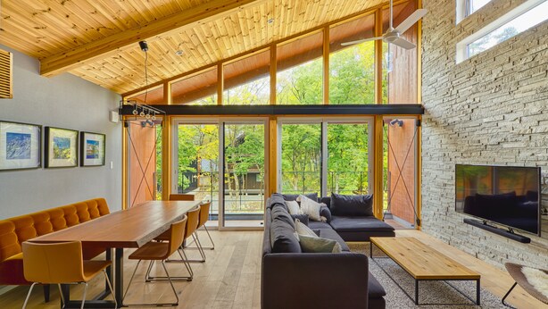 A modern living room with a wooden ceiling, large windows, a sofa, a TV, and a dining area. - Cherrywoods Place (Hakuba)