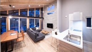 A modern kitchen with a built-in dishwasher, a dining area with a wooden table and chairs, a gray sofa, and a stone accent wall. - Cherrywoods Place (Hakuba)