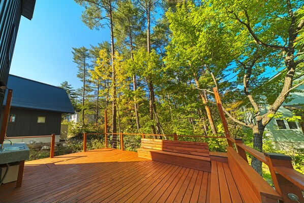 Una terraza de madera con barandilla metálica, rodeada de árboles y una casa.