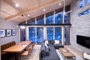 Una sala de estar moderna con techo de madera, ventanas grandes, chimenea y comedor.