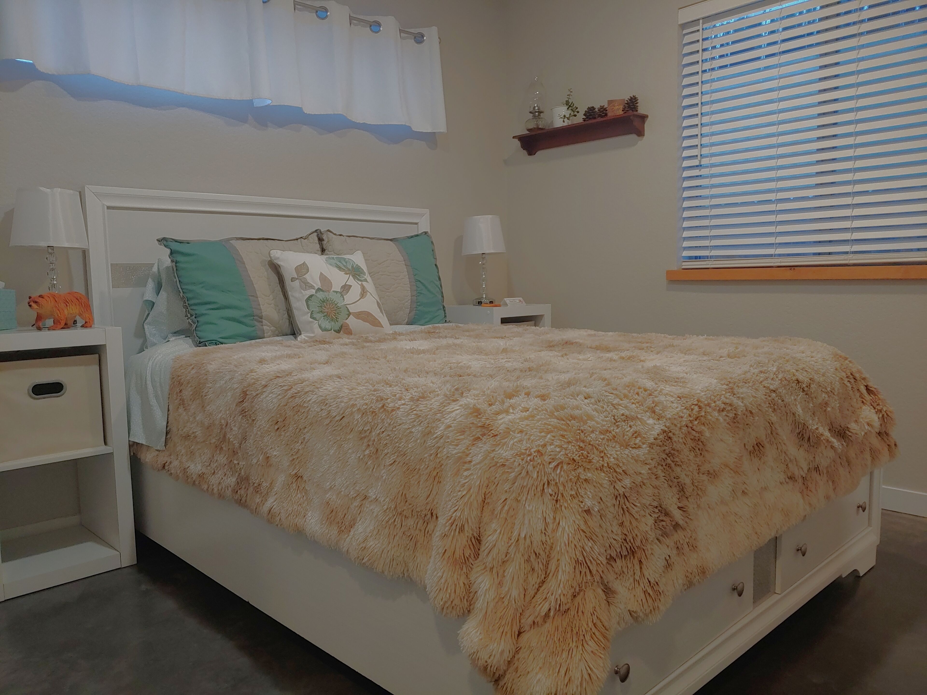 A neatly arranged bedroom with a large bed, two bedside tables, a nightstand, a wall-mounted shelf, and a window with blinds.