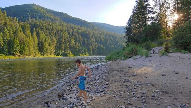 Une personne se tient au bord d’une rivière, avec une montagne boisée en arrière-plan.