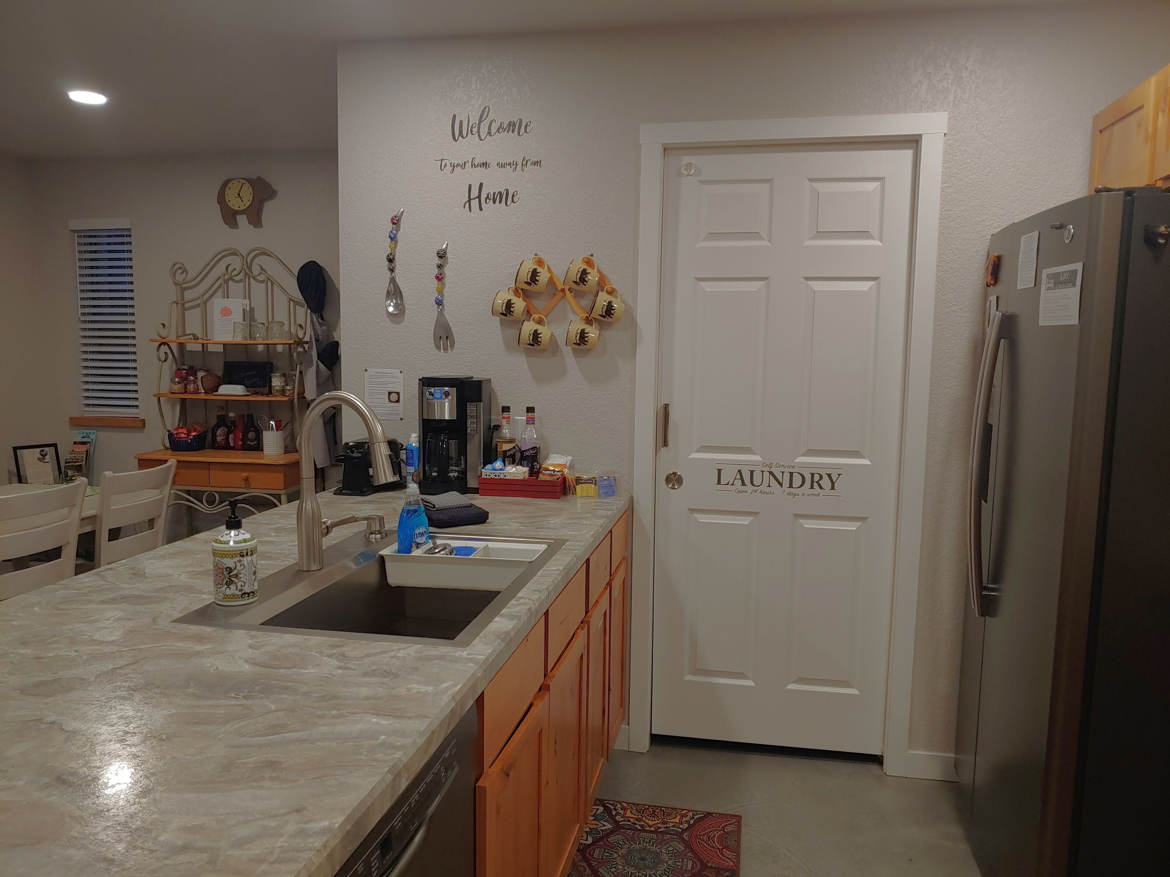 Een keuken met een marmeren aanrecht, een spoelbak, een koffiezetapparaat en een koelkast. Er is een deur met de tekst 'Laundry' en een muur met een welkomstbord.