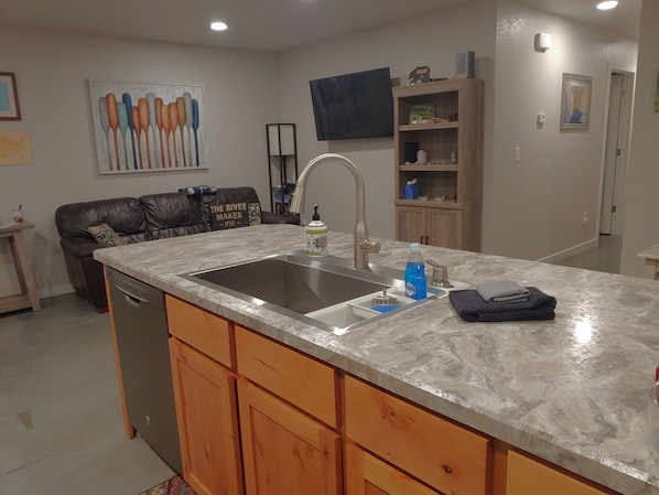 A modern kitchen with a large island, stainless steel sink, wooden cabinets, and a flat-screen TV mounted on the wall.