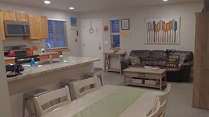 Il s’agit d’un espace de vie compact doté d’un coin cuisine comprenant un four à micro-ondes, une cuisinière et un évier. On y trouve une table à manger avec des chaises, un canapé et une table basse. La pièce est agrémentée d’une porte ornée d’une guirlande et d’un tableau représentant des cuillères accroché au mur.