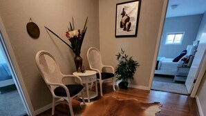 Una habitación con piso de madera, dos sillas blancas, una mesa con un florero y un cuadro en la pared, además de una puerta que da a otra habitación.
