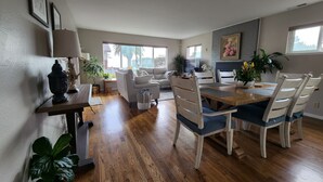 Amplia sala de estar con una mesa de comedor de madera, sillas blancas y un sofá. Se observa un televisor y una chimenea al fondo.