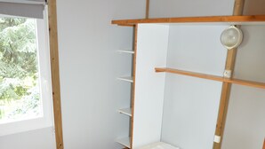 Una cama individual con estructura de madera, colchón blanco y colcha rosa. Se ve una ventana con persiana blanca, y una repisa con un cuenco blanco encima.