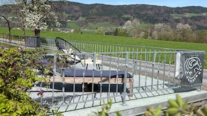 Eine Metallgeländer-Konstruktion mit einer Bank und einem Tisch, umgeben von Grünpflanzen und einer schönen Aussicht auf Hügel.