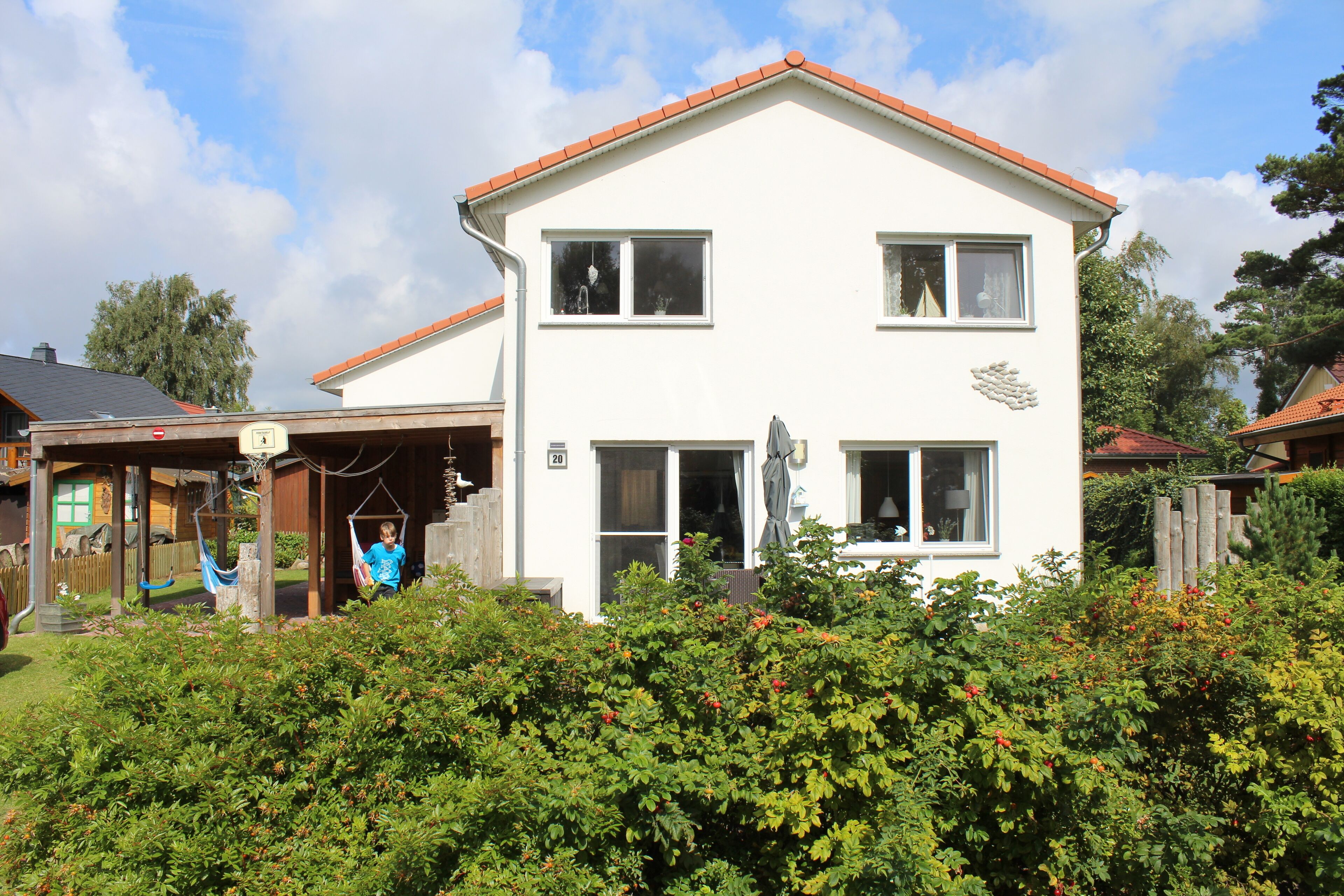 Ein zweistöckiges weißes Haus mit rotem Dach, einer Veranda und einem kleinen Garten mit Sträuchern und einer Hängematte.