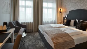 Et hotelværelse med seng, skrivebord med stol, sofa og et stort vindue med gardiner.