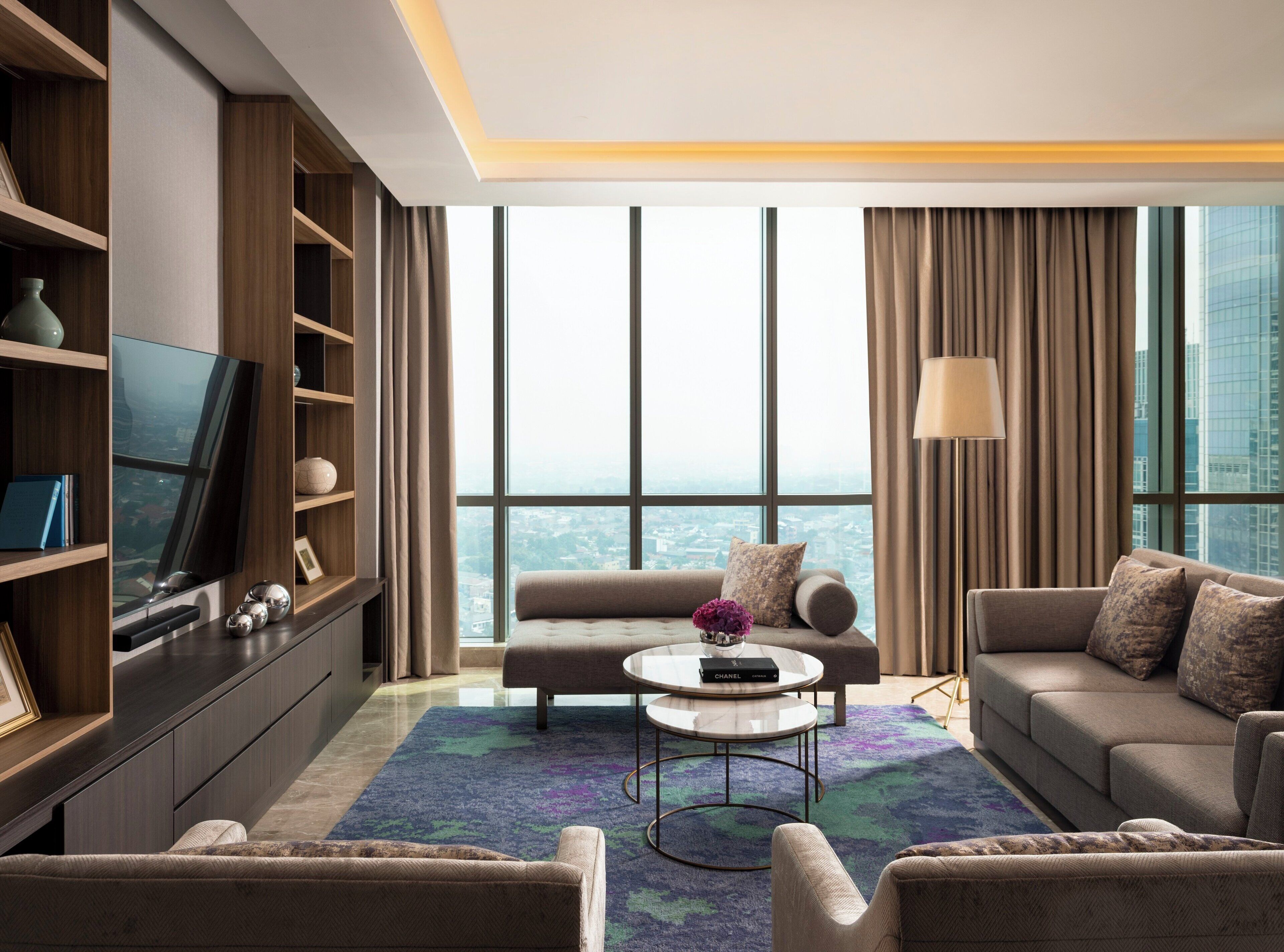 A modern living room with a large flat-screen TV, a sofa, a coffee table, and a view of a cityscape through large windows.