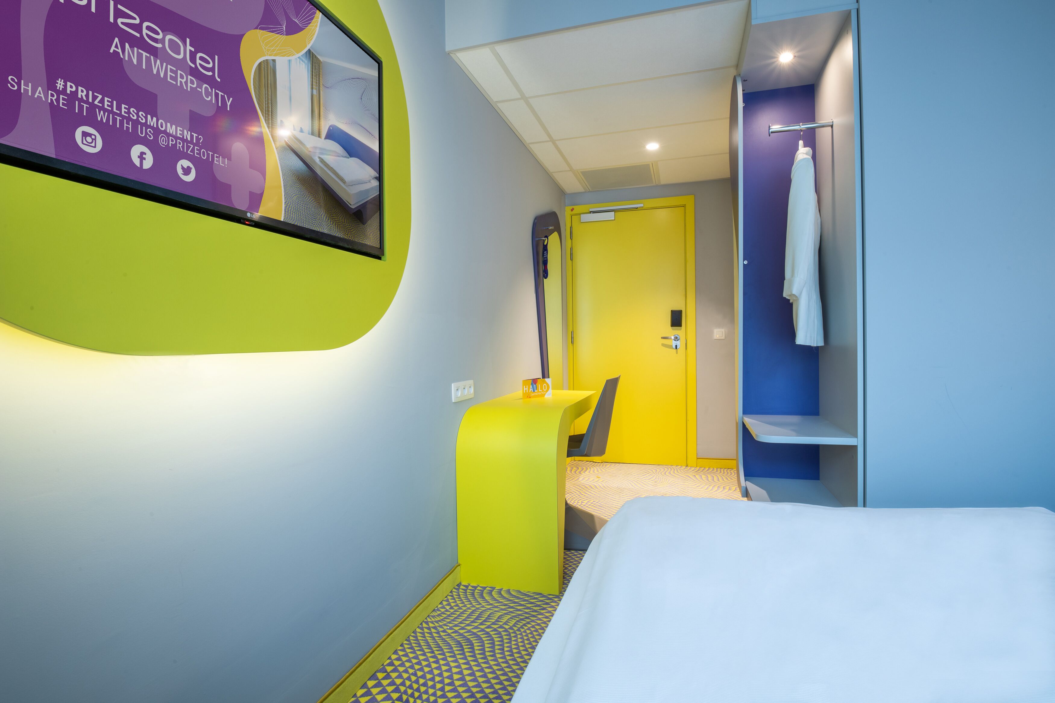 Hotelkamer met een bed, een spiegel, een klein bureau met stoel, een gele deur en een handdoekhouder.