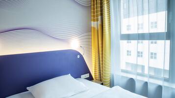 A hotel room with a modern design, featuring a blue headboard, a neatly made bed, a window with yellow curtains, and a view of buildings outside.