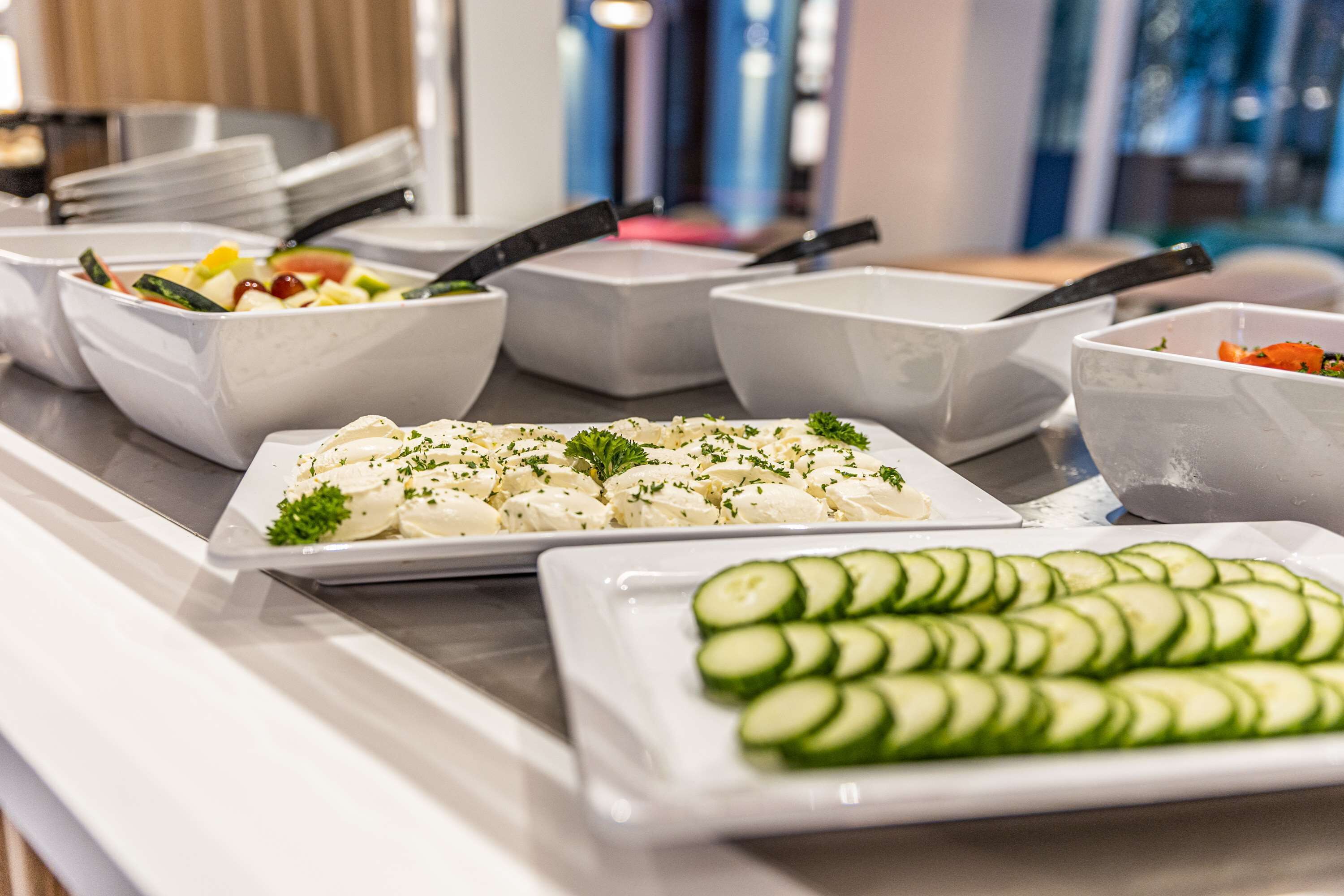 Un bufé con diversos platos, incluyendo un plato de pepinos en rodajas, una ensalada y un recipiente con una sustancia cremosa blanca adornada con hierbas.