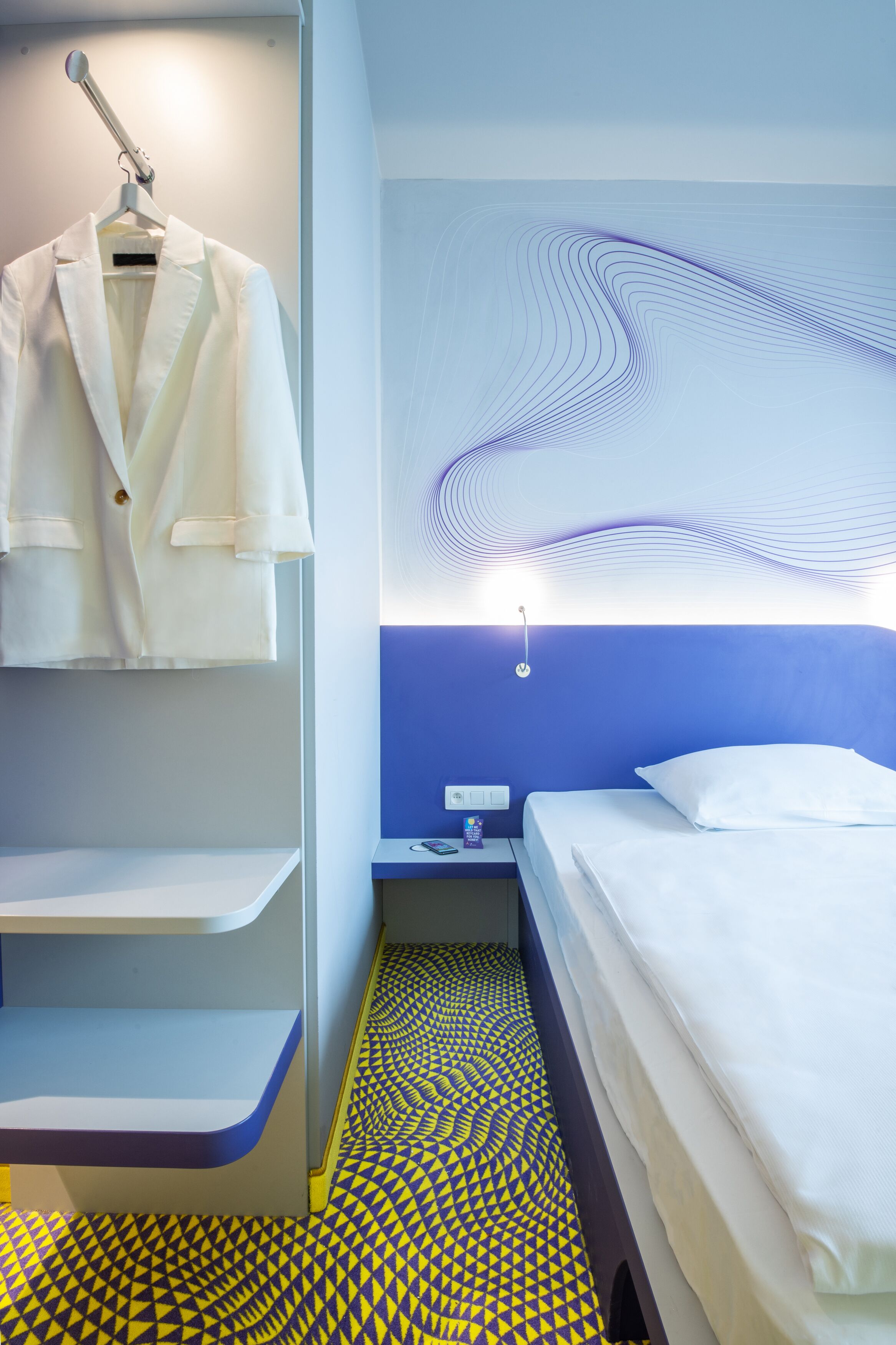 A hotel room with a bed, a nightstand, a robe hanging, and a patterned floor.