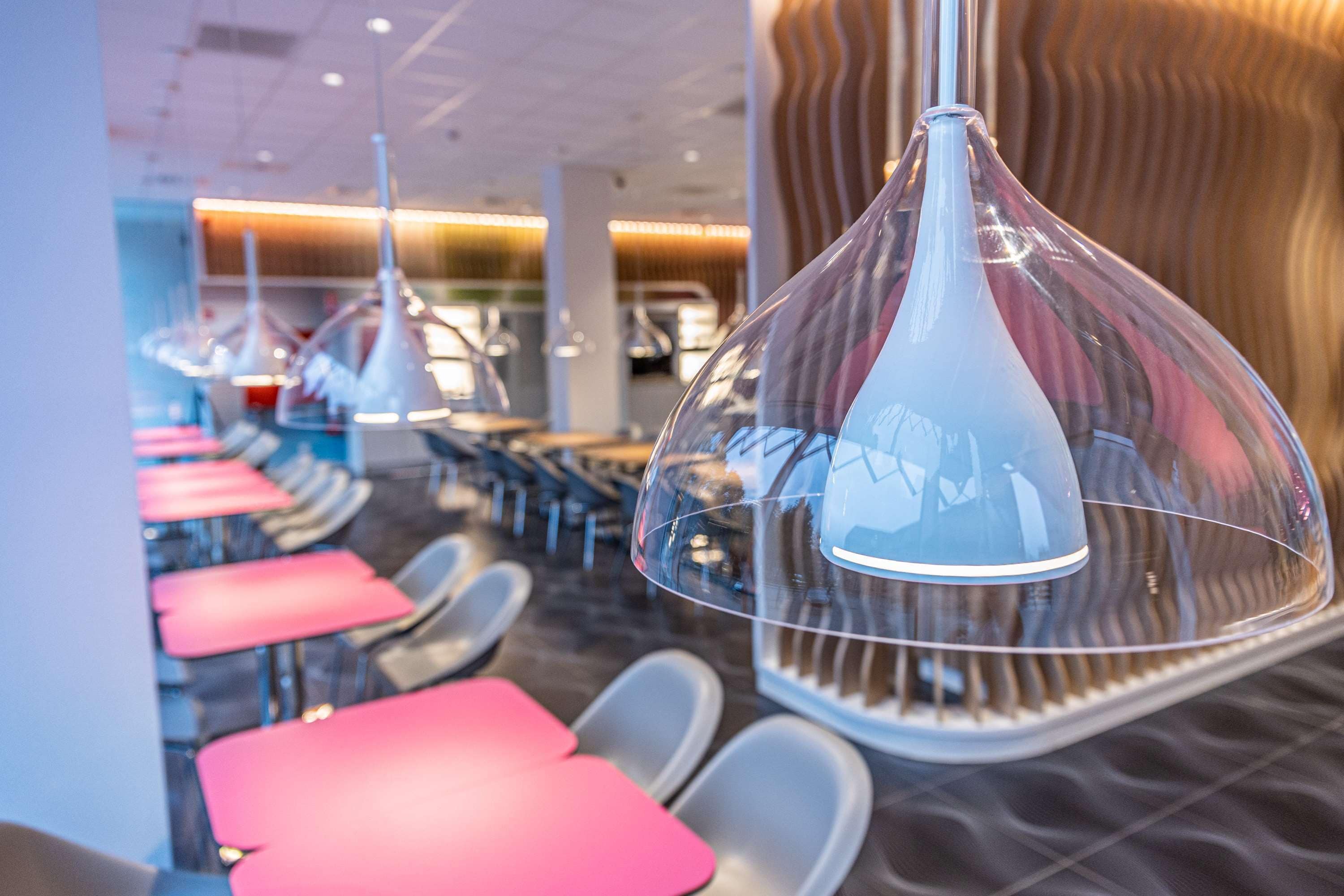 A modern restaurant interior with rows of tables and chairs, clear glass pendant lights, and a patterned wall.