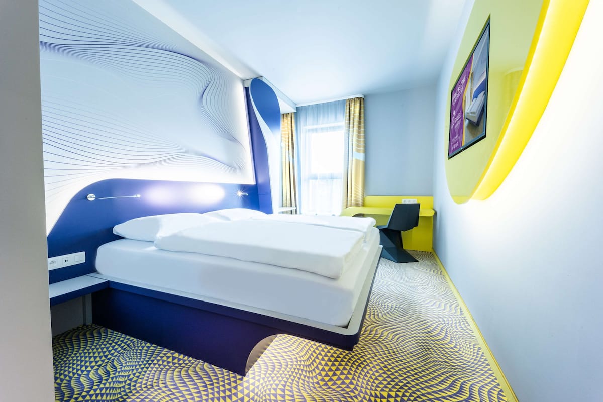 Ein modernes Hotelzimmer mit einem ordentlich bezogenen Bett, einem Schreibtisch mit Stuhl und einem großen Fenster mit Vorhängen.