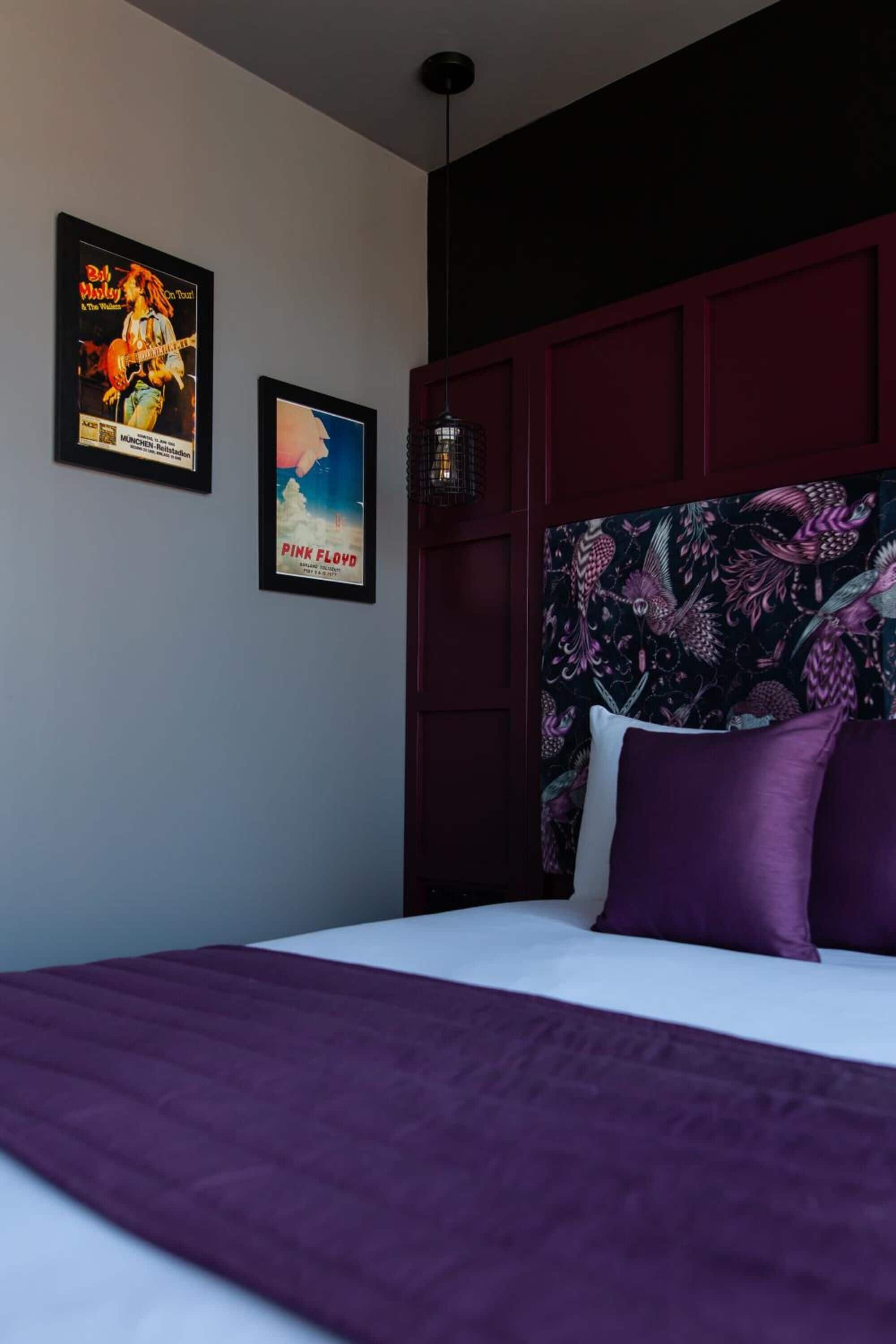 A hotel room with a large bed, a headboard with a floral pattern, and two posters on the wall.
