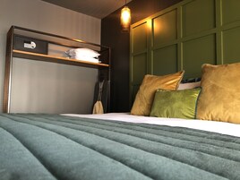 Una cama bien hecha con colcha verde, cabecero con estante y lámpara colgante.