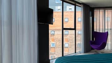 A modern hotel room with large windows, a bed, a purple chair, and a flat-screen TV.
