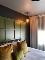Una habitación de hotel con una cama grande, cabecera con lámpara y ventana con cortinas.