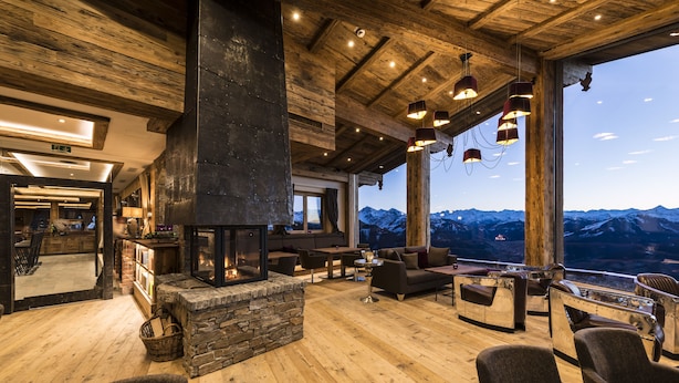A spacious lounge area with a stone fireplace, wooden flooring, and a view of snow-capped mountains.