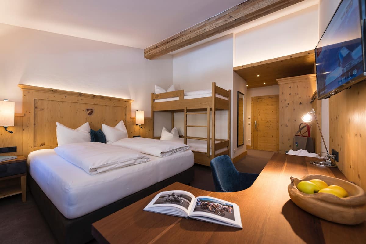 A hotel room with a large bed, a bunk bed, a desk with a magazine, and a fruit bowl.