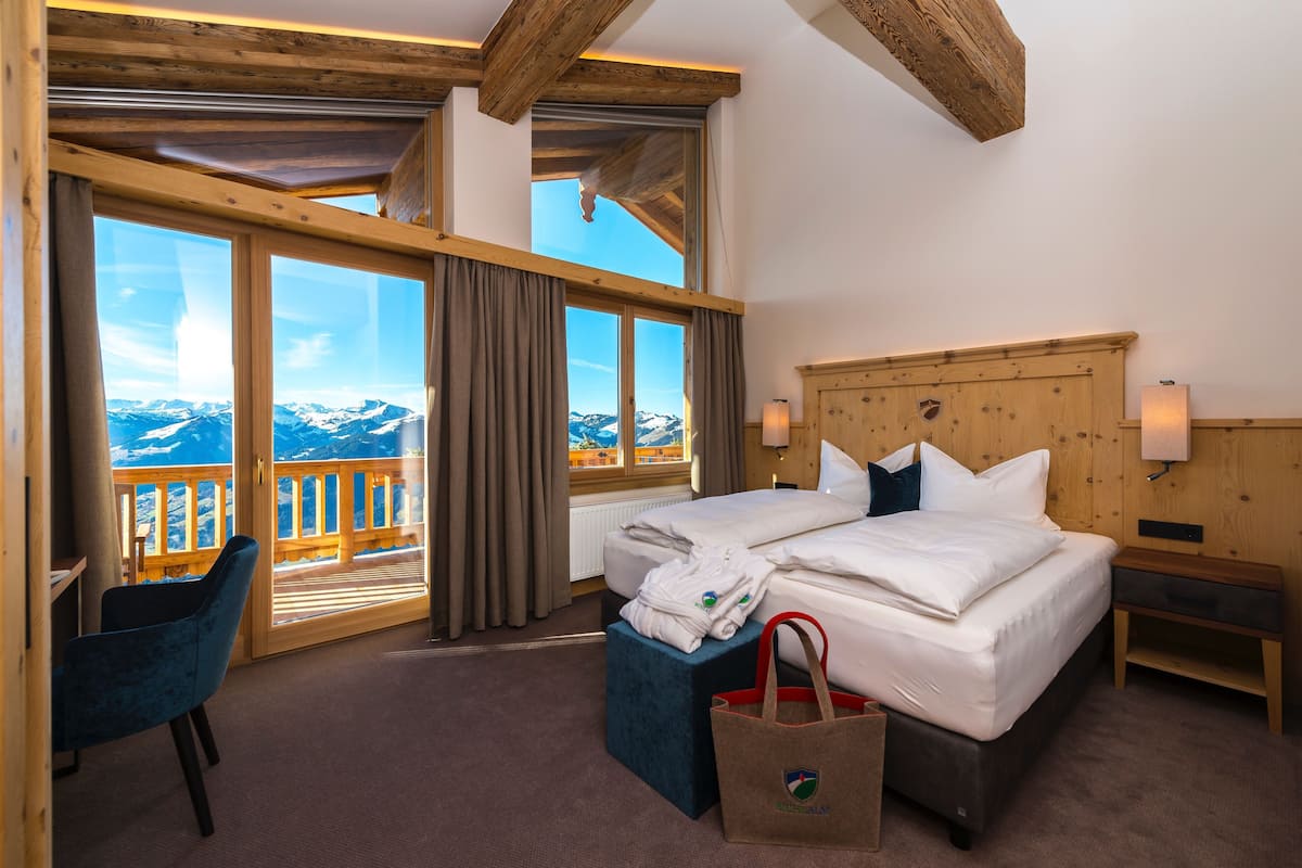 A hotel room with a wooden ceiling, a large bed, a view of mountains, and a wooden desk with a chair.