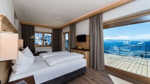 A modern hotel room with a large bed, a TV, a wooden desk, and a balcony with a mountain view.