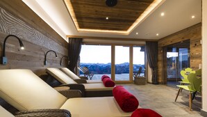A modern spa room with wooden paneling, reclining chairs, and a view of mountains through large windows.
