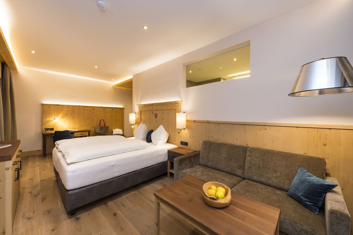 A hotel room with a bed, a sofa, a wooden coffee table with fruit, a desk with a lamp, and a mirror on the wall.