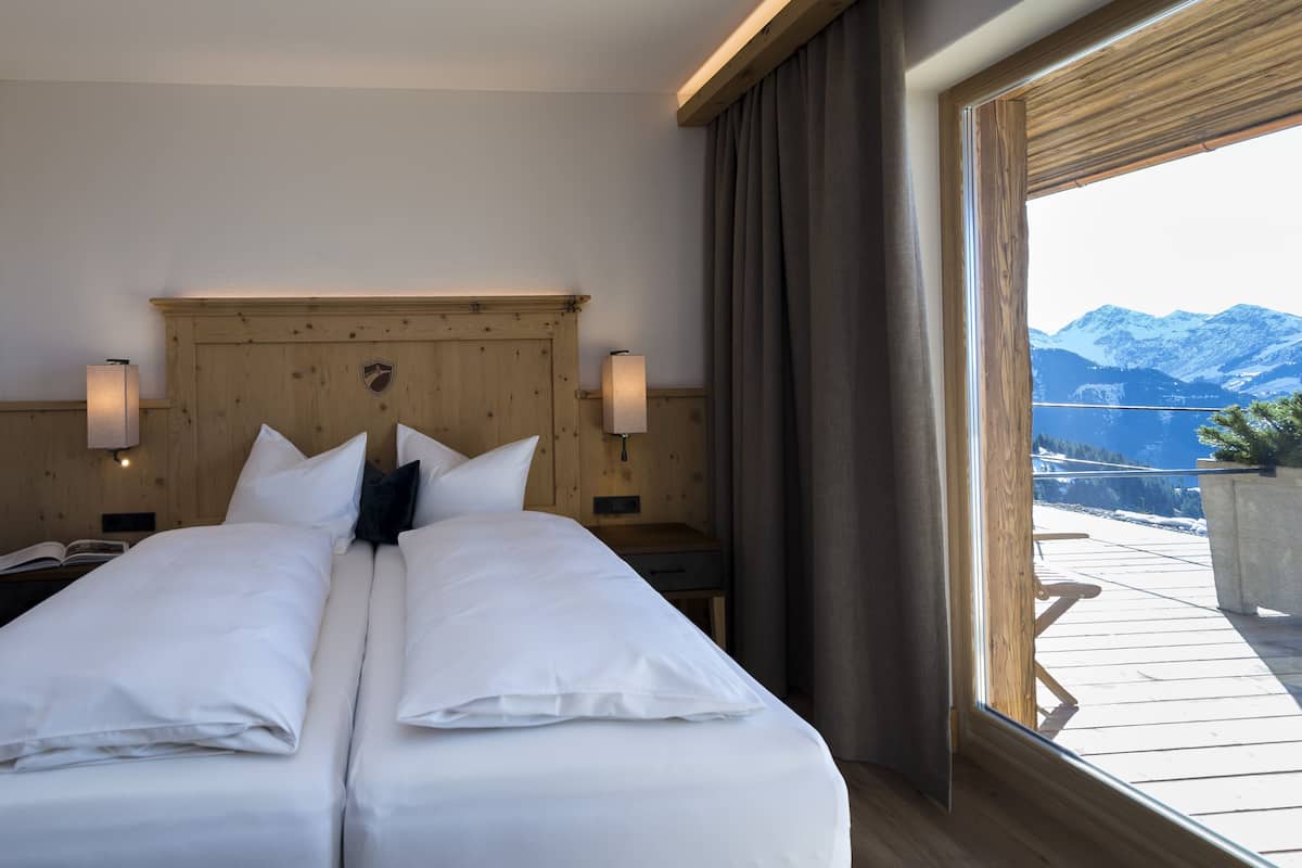 A double bed with white linens, wooden headboard, two bedside lamps, and a view of snow-capped mountains through a large window.