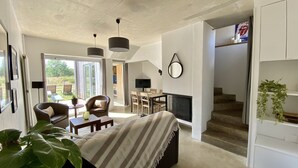 A modern living room with a sofa, dining table, and chairs. There is a fireplace, a TV, and a staircase leading to another level. - Les Gîtes de l'Ermitage (Terce)