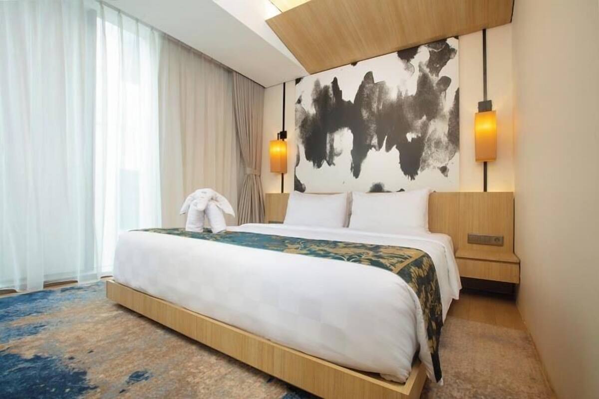 Quarto de hotel com uma cama grande, cabeceira com pintura abstrata preta, duas luminárias de cabeceira e uma janela ampla com cortinas translúcidas.