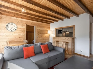 A modern living room with a wooden ceiling, a grey sofa with red cushions, a wooden bar counter, and a kitchen area with a microwave.