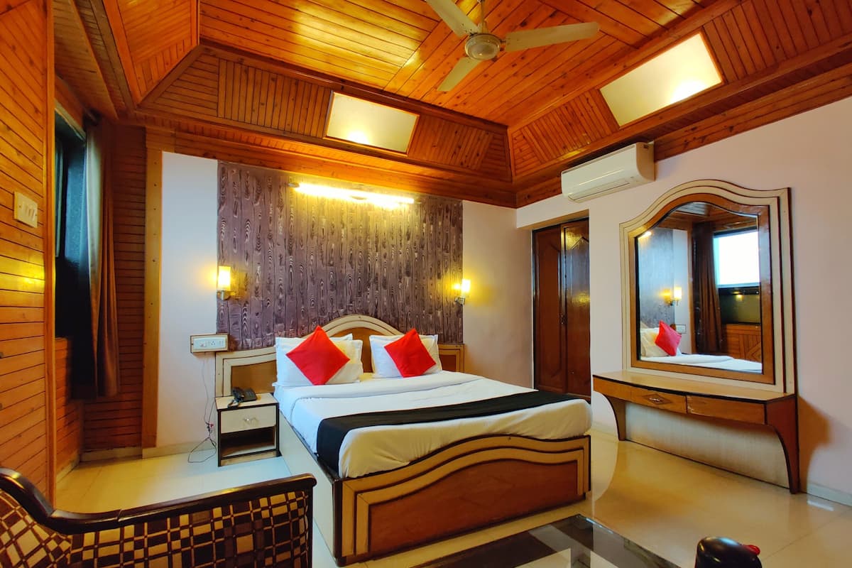 Habitación de hotel con techo de madera, cama con almohadas rojas y blancas, mesita de noche con lámpara, una silla y un espejo grande.