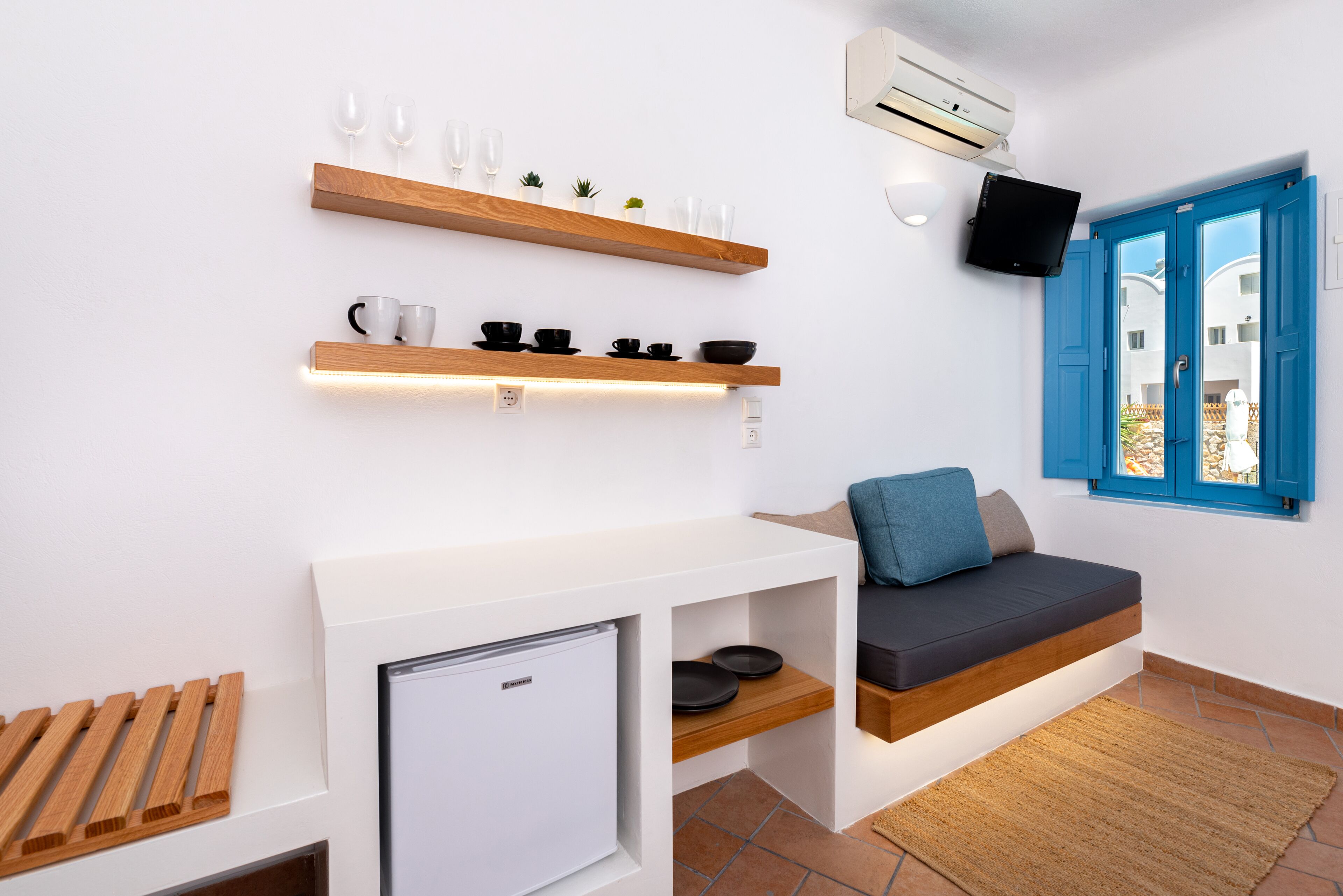 Un espace de vie compact comprenant un coin cuisine, un espace détente avec un canapé et des coussins, et une fenêtre avec des volets bleus.